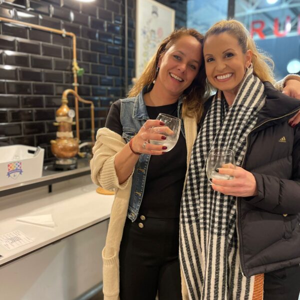 two gin school students standing in front of a Gin School classroom holding a glass of the gin they made