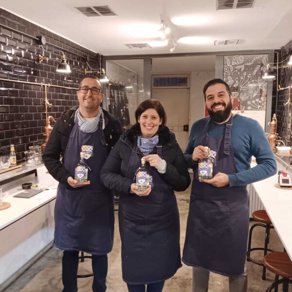 three gin school students standing in front of a Gin School classroom holding bottles of the gin they made